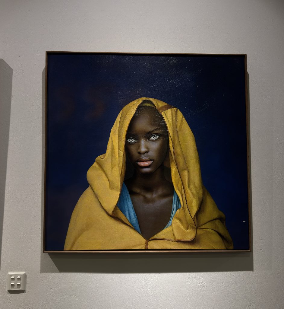 Woman in a yellow hood on a deep blue ground, framed on a white wall